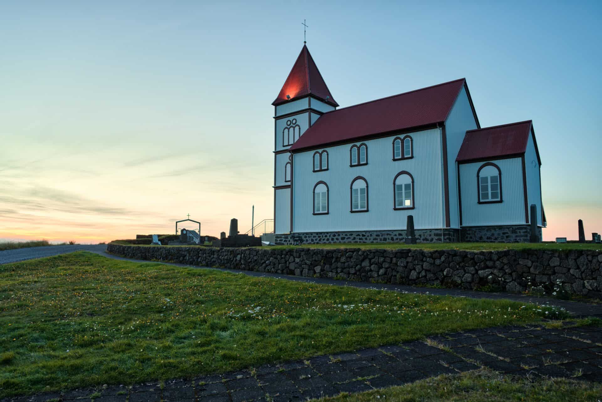 Scenic church with red roof and white walls, set against a colorful sunset sky, surrounded by green grass and stone wall.